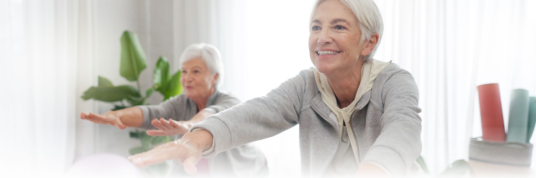 Two elderly women staying active, smiling and extending their arms forward. The logo for The Ohio Masonic Communities is visible above them. The softly lit background features plants and exercise mats, showcasing the vibrant atmosphere of this senior living community.