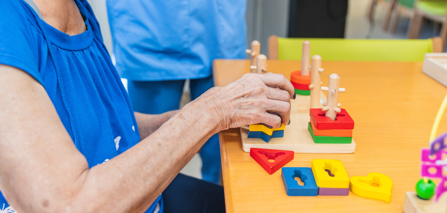 Older adult places blocks of different shapes in a puzzle with a medical professional standing by
