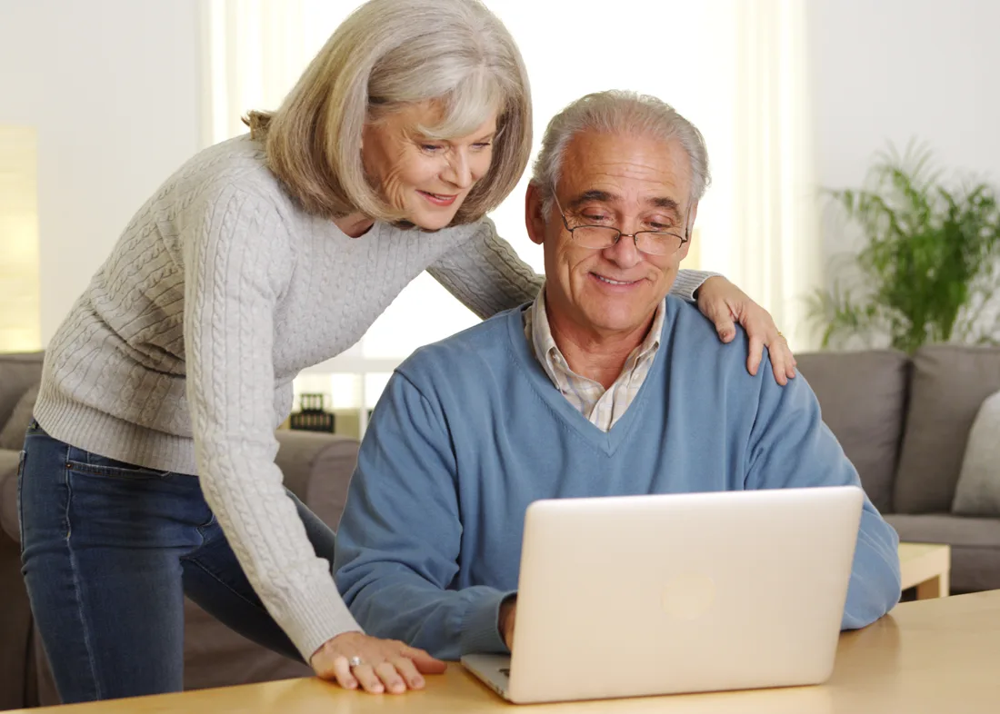 Older adult female leaning over older adult male as they look at a laptop together