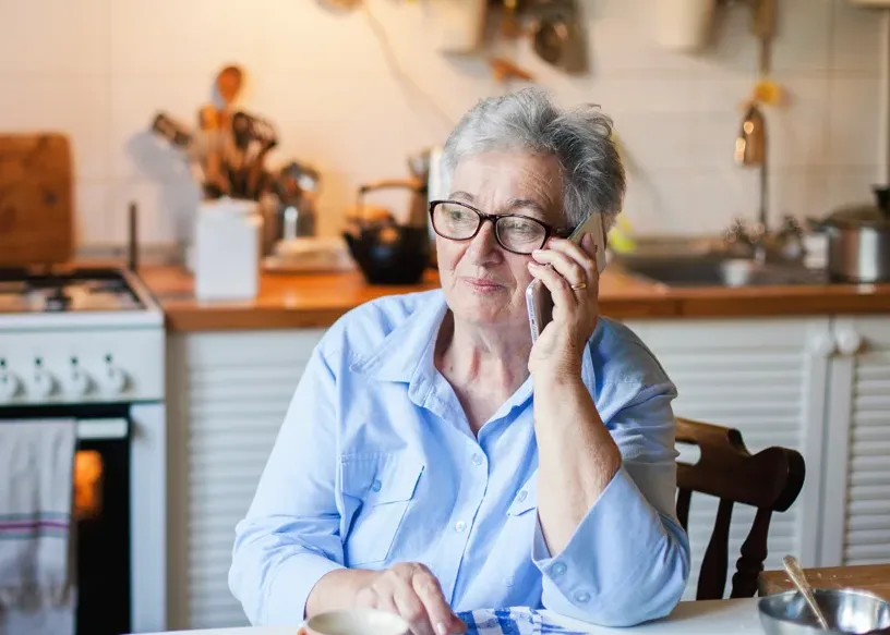 Older adult female sits in the kitchen and talks on her cell phone