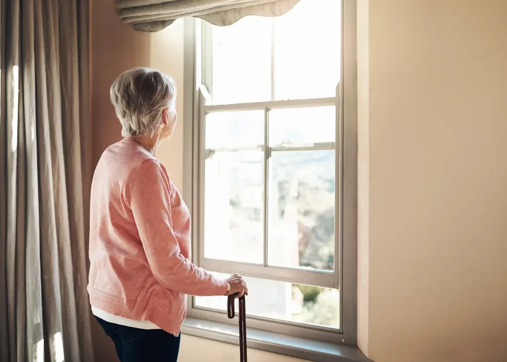 Senior woman stands with a cane and looks out a window